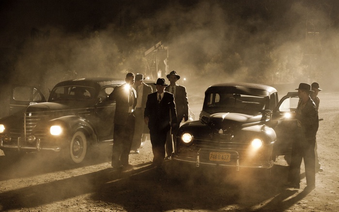 Men in suits and hats stand by classic cars at night