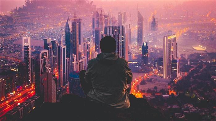 Man sitting on a cliff overlooking a city at dusk