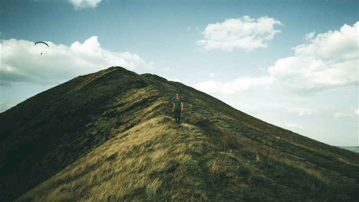 Man walking on a grassy mountain ridge with paraglider in the sky