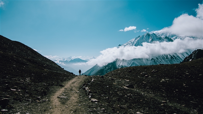 Man hiking on a mountain trail with clouds and snowy peaks