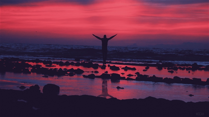 Silhouette of a person with arms outstretched on a rocky shore at sunset