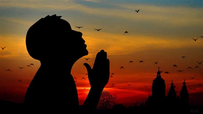 Silhouette of a person praying against a sunset sky with birds and a church