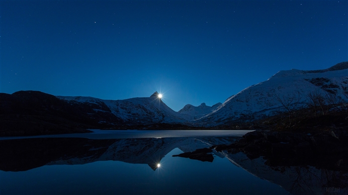Moon behind snow-capped mountains over a lake
