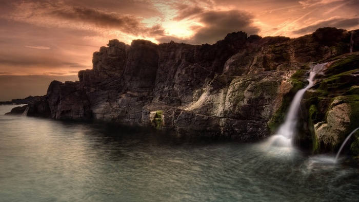 Waterfall cascading over mossy rocks into the sea at sunset