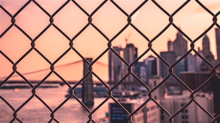 Chain-link fence with blurred city skyline and bridge at sunset