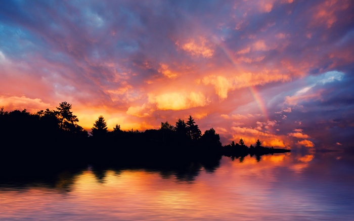 Sunset over a lake with a rainbow and silhouetted trees