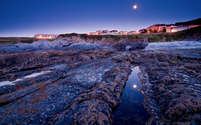 Rocky coastline at dusk with moon and town lights