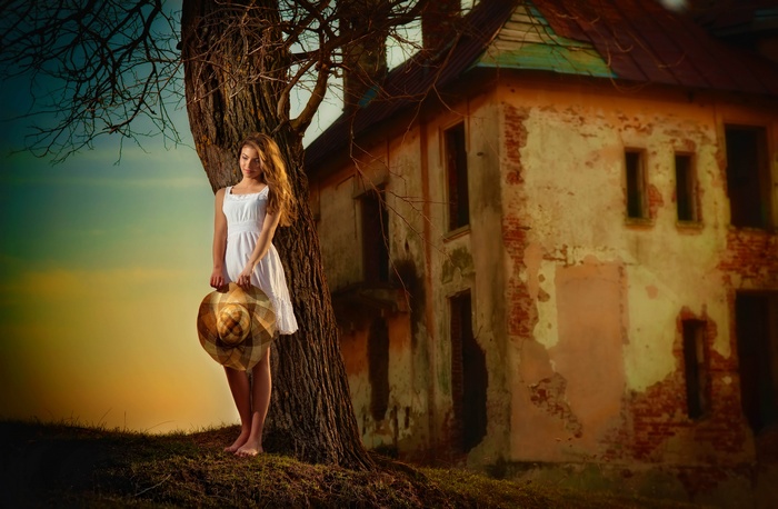 Woman in white dress holding a hat by a tree and old house