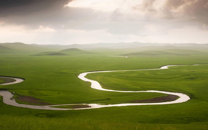 Winding river through green rolling hills under cloudy sky