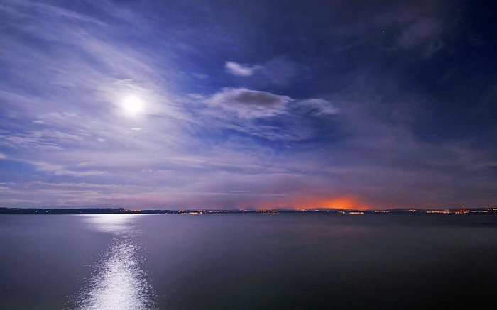 Moonlit lake with distant city lights under cloudy sky