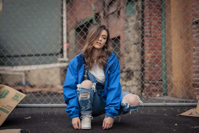 Woman in blue jacket and torn overalls crouching outdoors