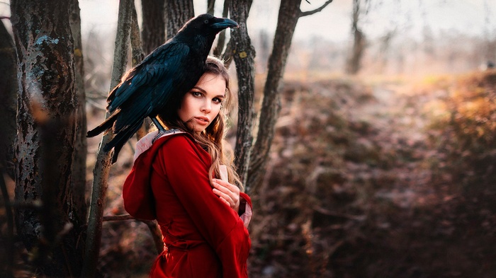 Woman in red jacket with raven on shoulder in forest