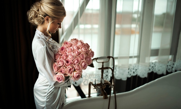 Bride in white robe holding pink roses by the window