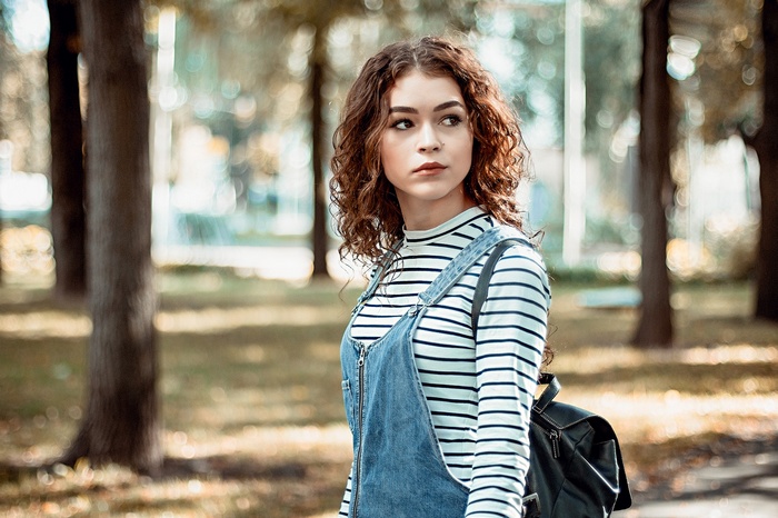 Young woman with curly hair wearing overalls and backpack outdoors