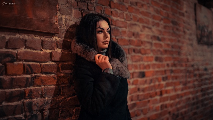 Woman in black coat with fur collar leaning against brick wall