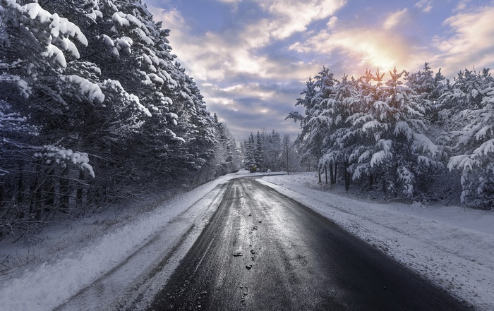 A snowy road winds through a winter forest under a cloudy sky.