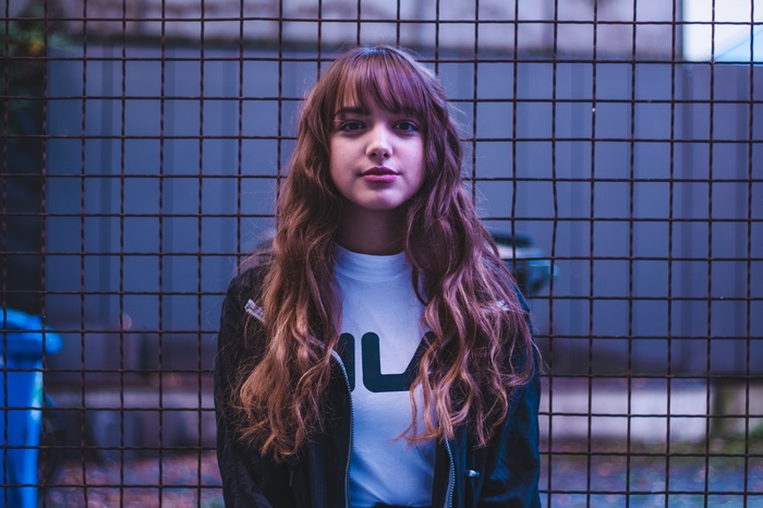 Woman with long brown hair and bangs standing in front of a fence