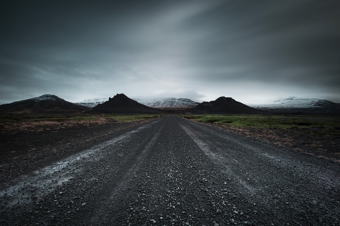 Gravel road leading to dark mountains under cloudy sky