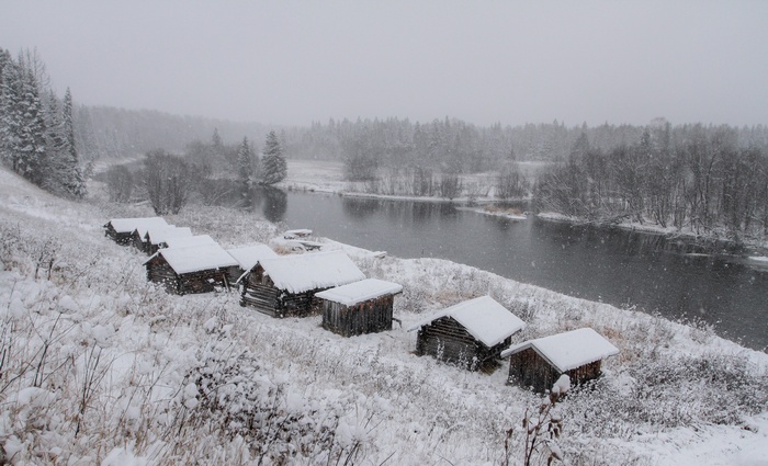 Wooden cabins by a river during a snowfall