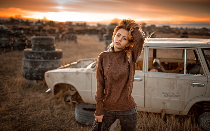 Woman in sweater posing near abandoned car at sunset