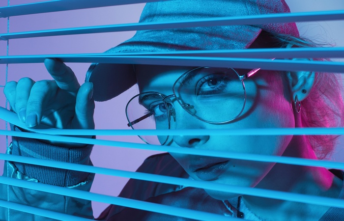 Woman in glasses and cap peeking through blinds