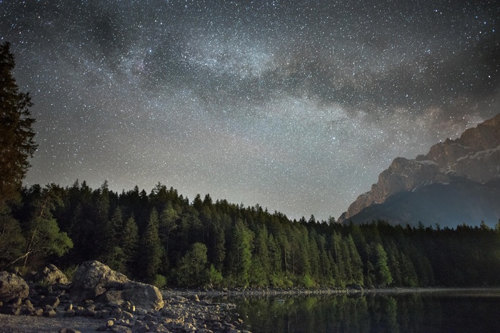 Starry night over forest and mountains reflected in lake