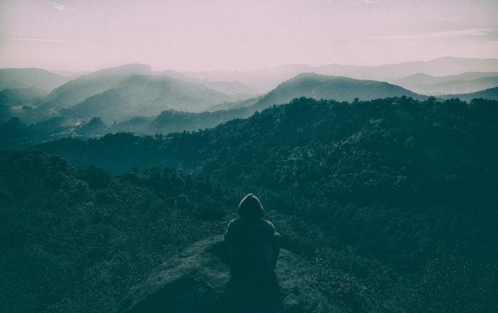 Person in hoodie sitting on a rock overlooking misty mountains