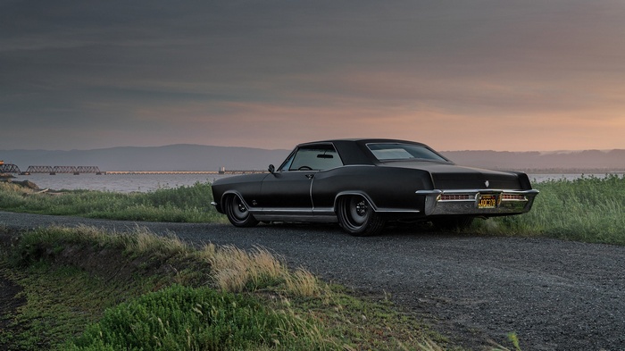 Black Buick Riviera on a gravel road near a bridge