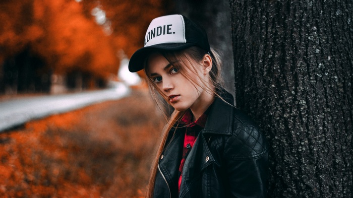 Woman in leather jacket and baseball cap leaning against tree