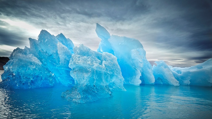 Blue iceberg floating in bright blue water under cloudy sky