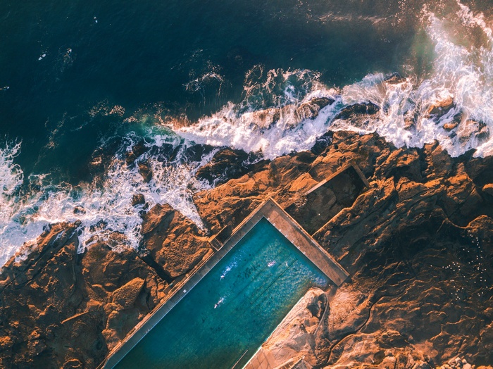 Aerial view of ocean waves crashing on rocks near a swimming pool