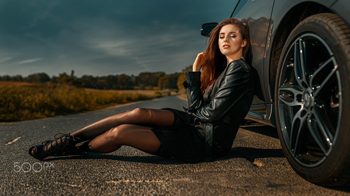 Woman in leather jacket leaning on car on a road