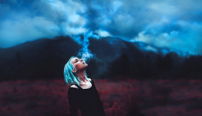Woman with blue hair exhaling smoke in a dark landscape