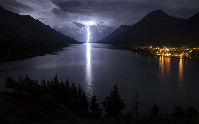 Lightning strikes over a lake with mountains and a town at night