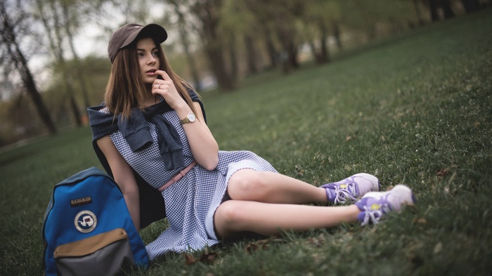 Woman in dress and baseball cap sitting on grass with backpack