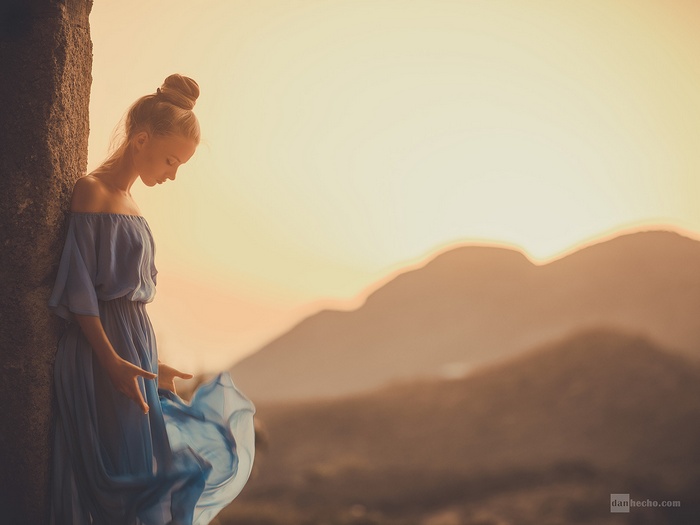 Woman in blue dress leaning against stone at sunset