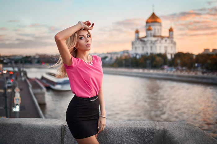 Woman in pink top and black skirt by river with church