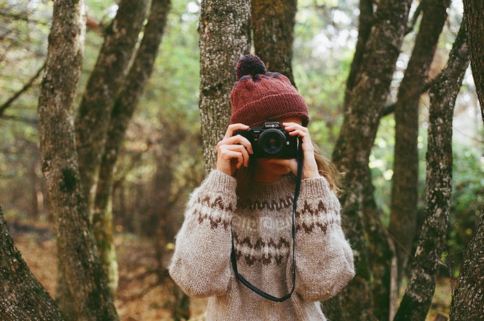 Woman in knitted sweater and hat taking photo with Minolta camera in forest