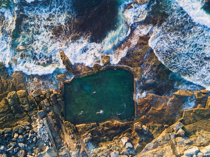 Aerial view of ocean pool with swimmers and waves crashing on rocks