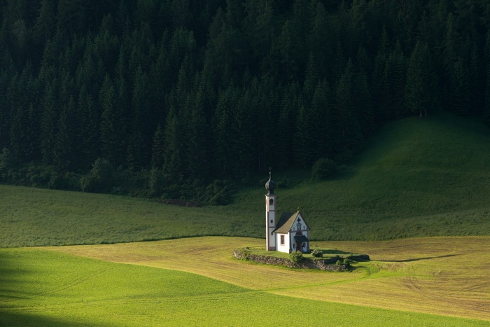 A small church stands alone in a green field with a forest behind it.