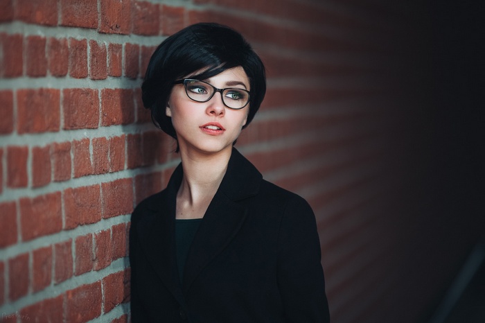 Woman with black hair and glasses leaning against a brick wall