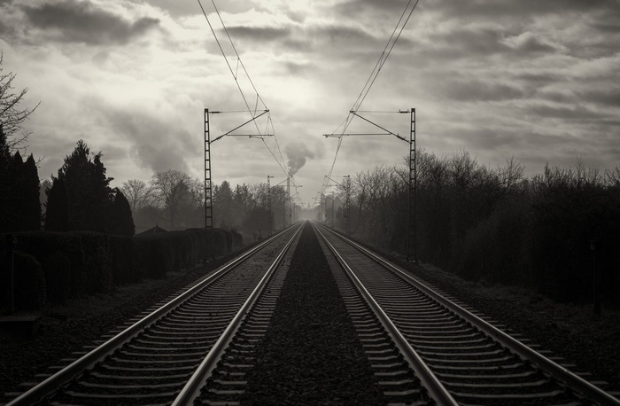 Railway tracks leading to a smoking industrial chimney under cloudy sky