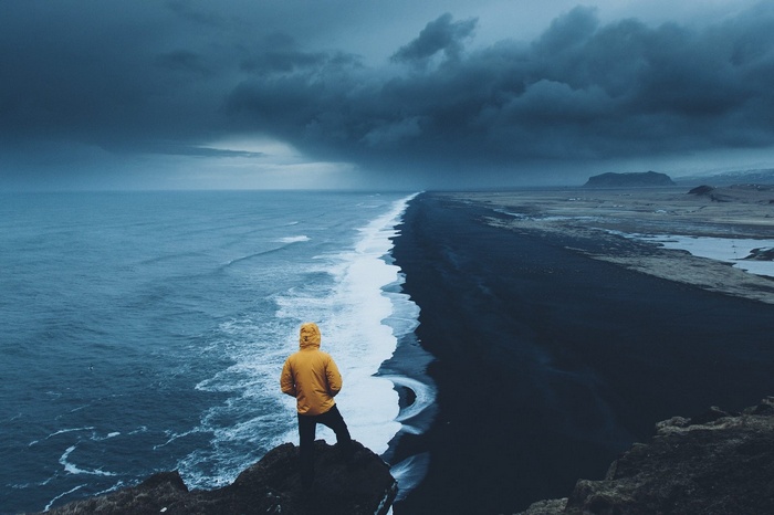 Person in yellow jacket on cliff overlooking black sand beach and ocean