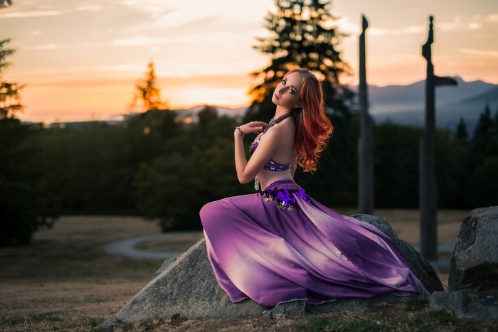Woman in purple belly dancing costume sitting on rock at sunset