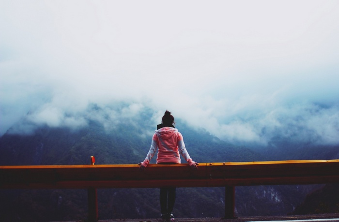 Woman in hoodie sitting on guardrail overlooking foggy mountains