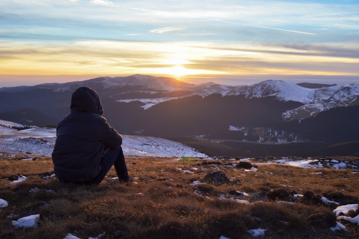 Person in Hood Sitting on Mountain Overlooking Snowy Landscape at Sunrise