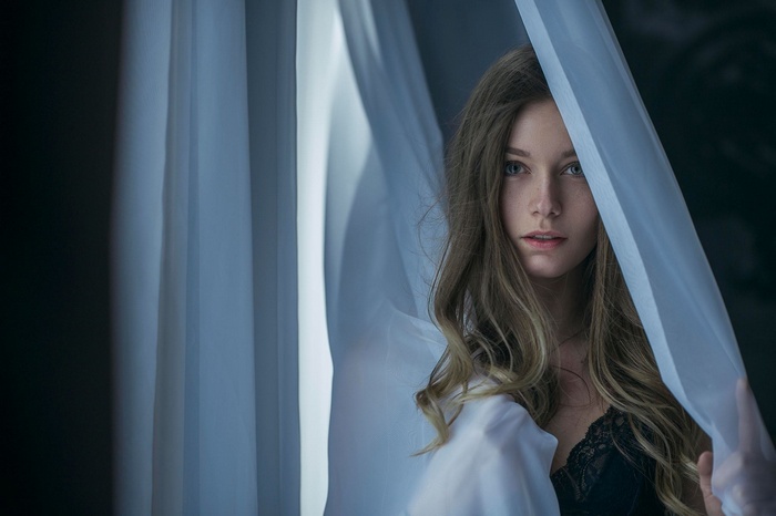 Woman with blonde wavy hair behind sheer curtains