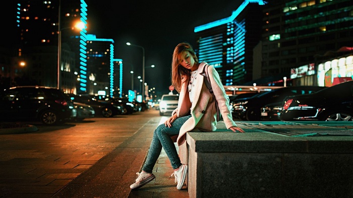 Woman in pink coat and jeans sitting on a ledge at night