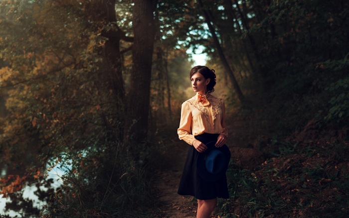 Woman in a peach shirt and black skirt holding a hat in a forest
