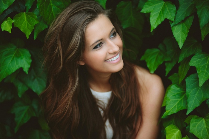 Woman with long wavy brown hair smiling among green leaves